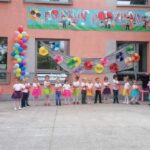 Zdjęcie przedstawia dzieci z grupy Biedronki podczas występu dla rodziców. Stoją na tarasie ustawione w półkolu. Ubrane są w kolorowe stroje, w rękach trzymają czerwone tasiemki.